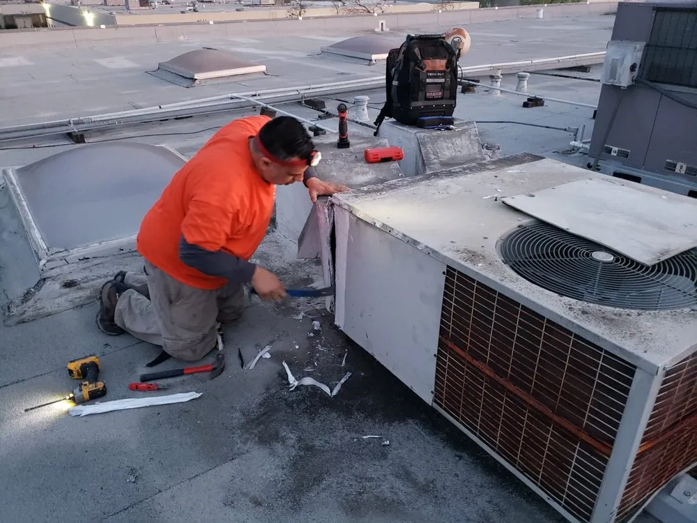A technician in an orange shirt working on an HVAC unit on a rooftop, surrounded by tools, representing InsulationGuruInc.dbaHVACGuru's dedication in SanJose.